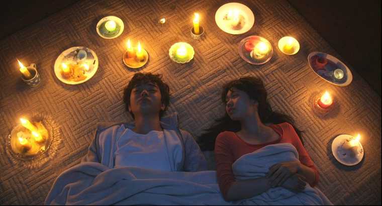 A teenage boy and girl lie clothed under a blanket, surrounded by lit candles