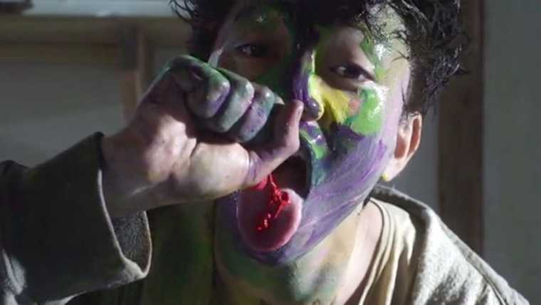 A teenage boy whose face is covered in paint, squeezes red paint on to his tongue