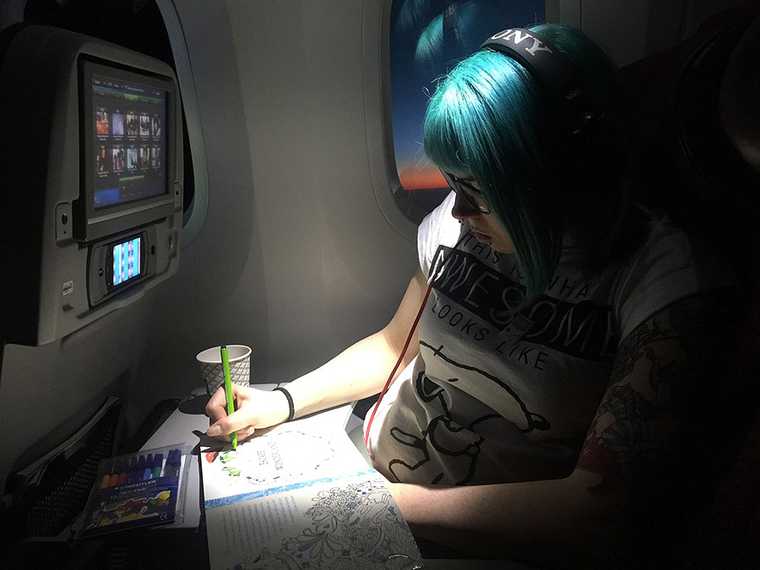 Blue haired girl sitting in a plane seat. She is wearing headphones and colouring-in a picture book.