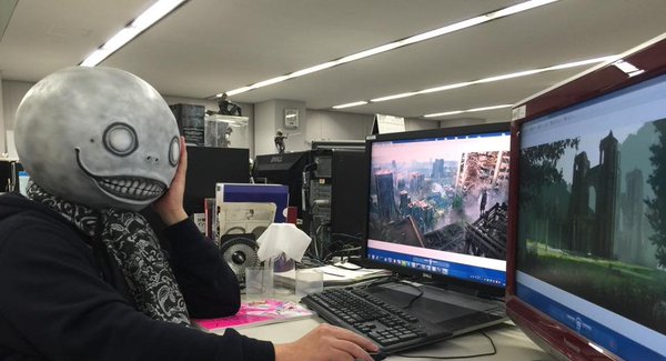 A man wearing a creepy mask (Yoko Taro) sits at a desk with two computer screens.