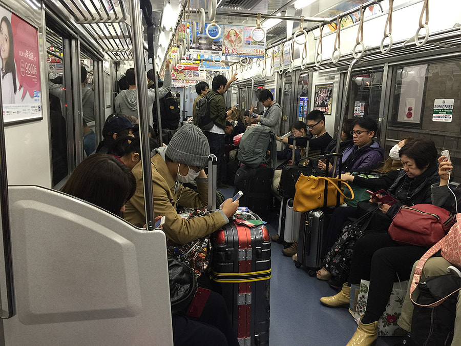 A Japanese train carriage with many people and suitcases. Most of the passengers are using their mobile devices.