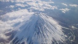 Photograph of Mt Fuji from above. The peak is covered in snow, and there are clouds on the far side.