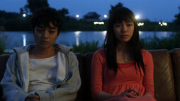 A teenage boy and girl sit on a sofa in front of a river