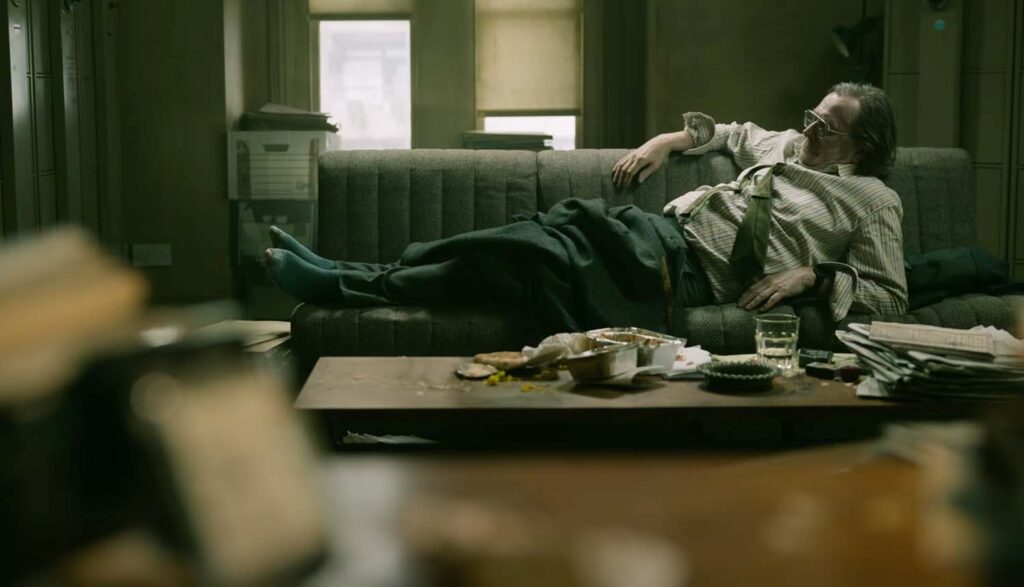 Screen from Slow Horses TV show. Gary Oldman as Jackson Lamb, reclined on a sofa. The room is littered with food containers and paperwork.