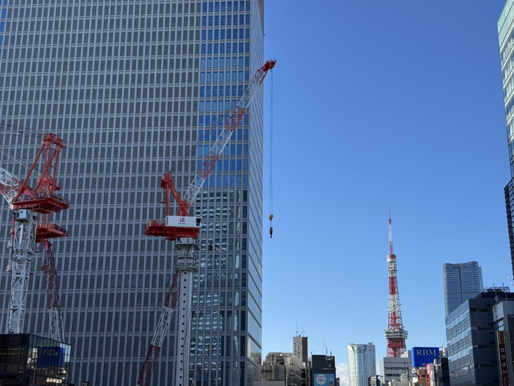 A tall building, and two cranes in front of it. I the back, Tokyo Tower against a blue sky.