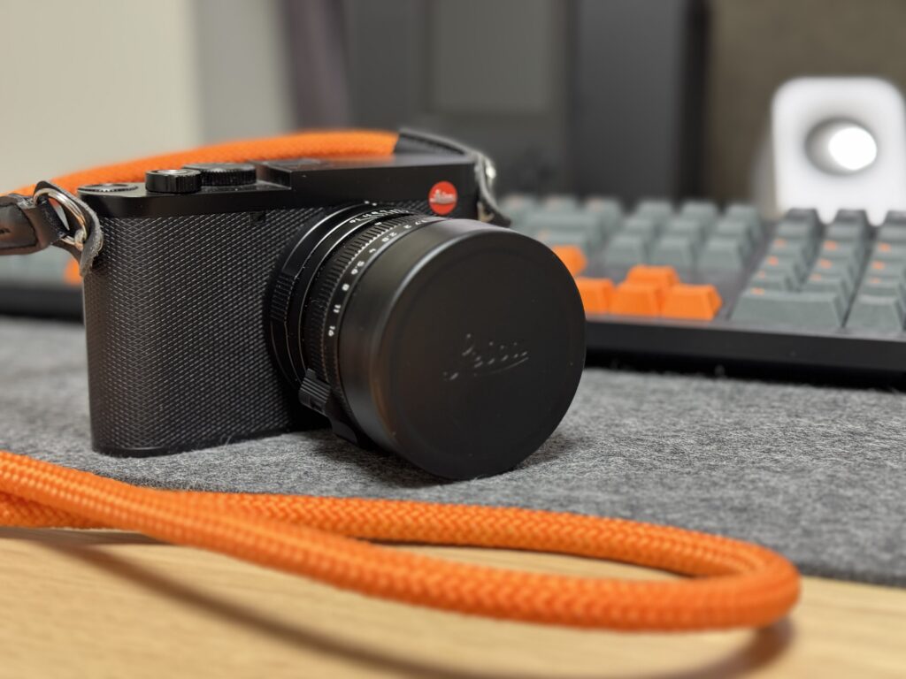 Leica Q3 camera with an orange rope strap sitting on a desk facing the camera. In the background an out of focus keyboard and speaker.