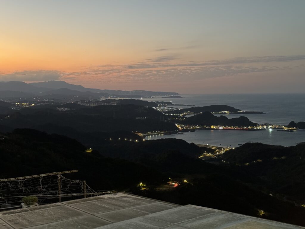 Jioufen from above - looking at the sea during dusk. Dark mountains, and lights scattered throughout.