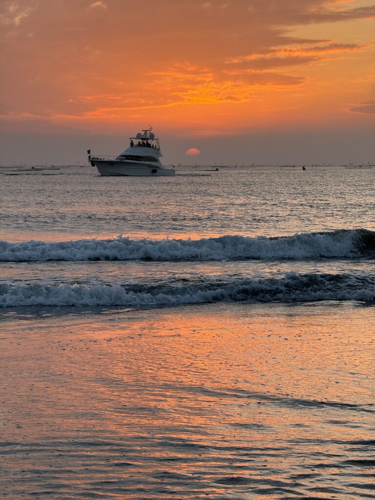 Yuguang beach at sun set. The sky is orange, and the sun dim on the horizon. A single yacht passes.