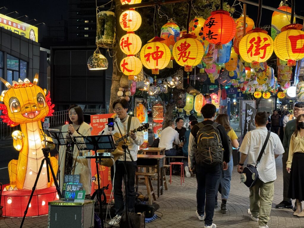 A night market in Tainan. A couple play music, the man playing guitar, under various coloured lanterns.