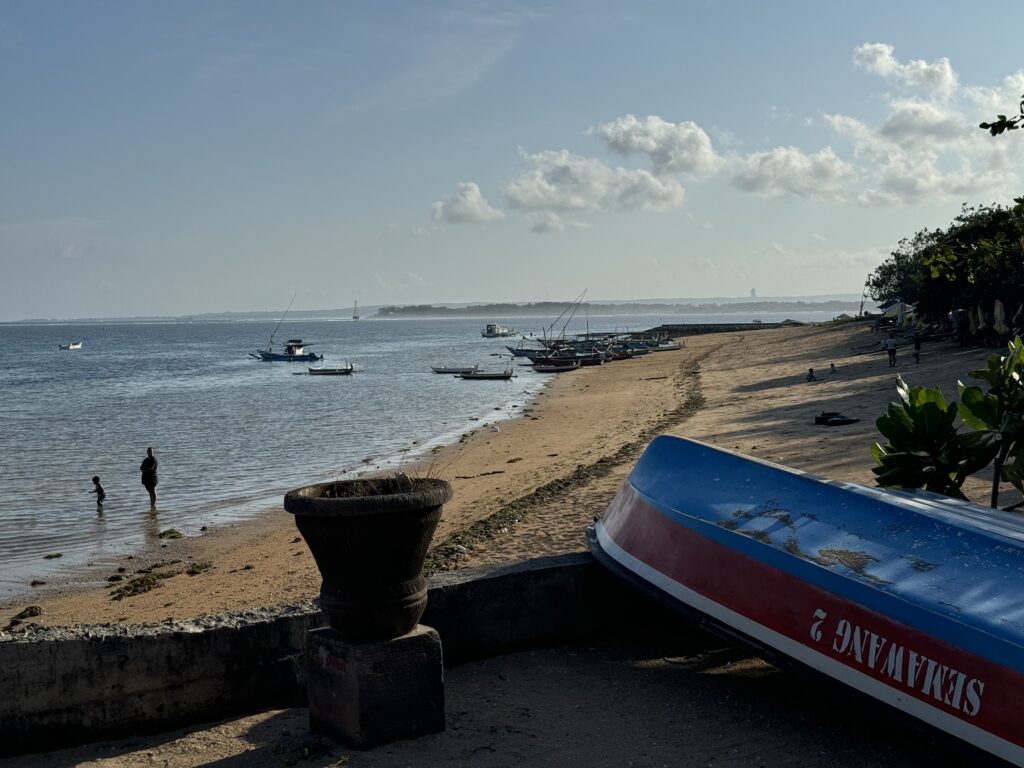 The beach and ocean in Bali. A mother and child stand in the shallow water. In the background, multiple boats line the coast. In the foreground, an upturned boat.