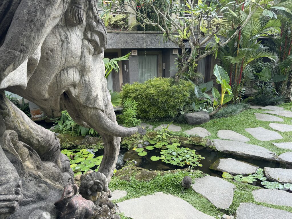 A hotel courtyard in Bali. A stone elephant statue in the foreground, appears to look into a garden area, with water feature and lots of foliage.