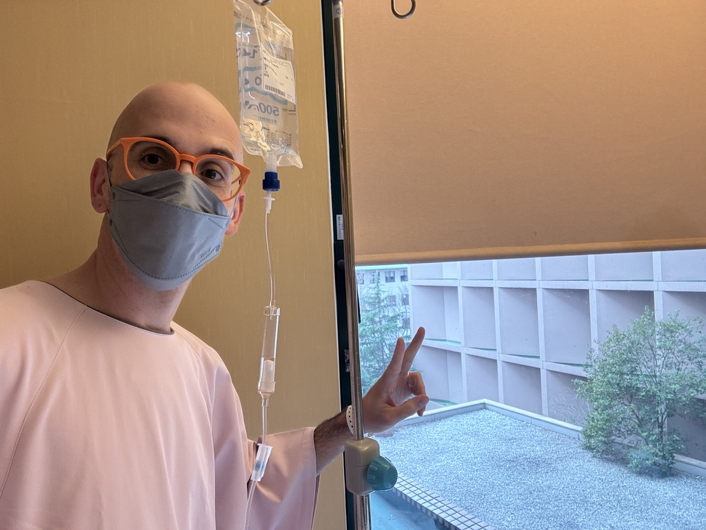 Bald middle aged man wearing a mask and glasses. He is attached to an IV drip and standing by a window.